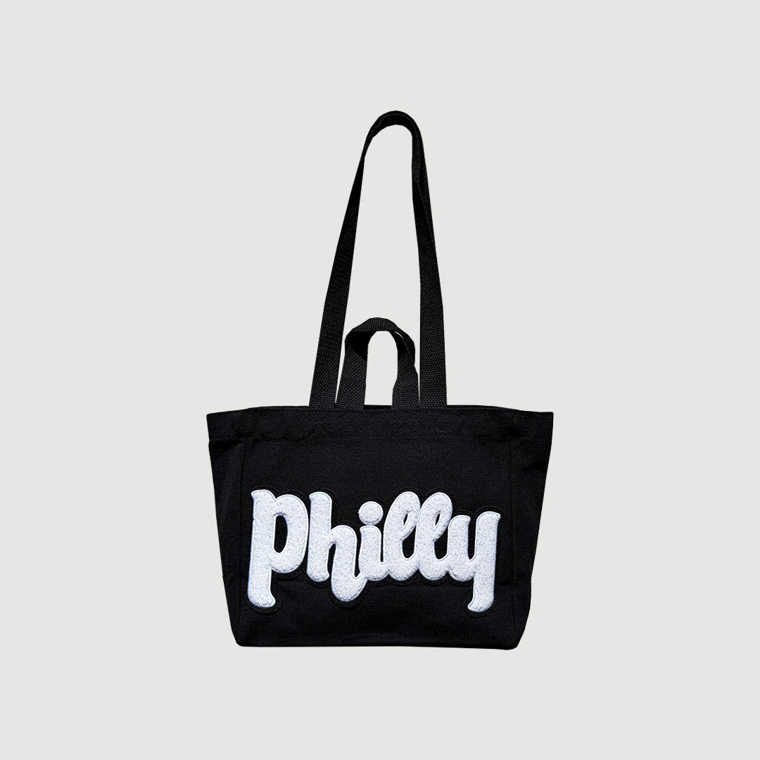 A black canvas tote bag with long shoulder straps and a top carry handle, featuring the word 'Philly' in large white bubble letters across the front.