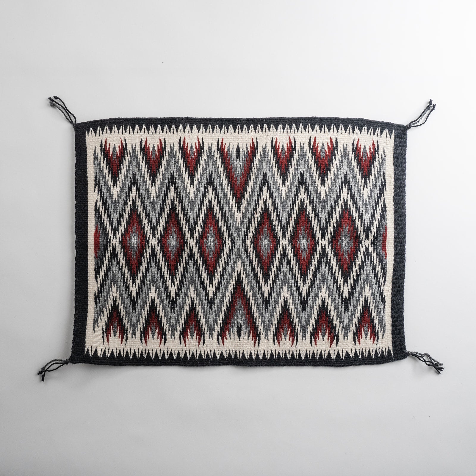 A rectangular woven rug with a Navajo eye dazzler geometric pattern in black, off-white, and dark red, featuring fringed corners.