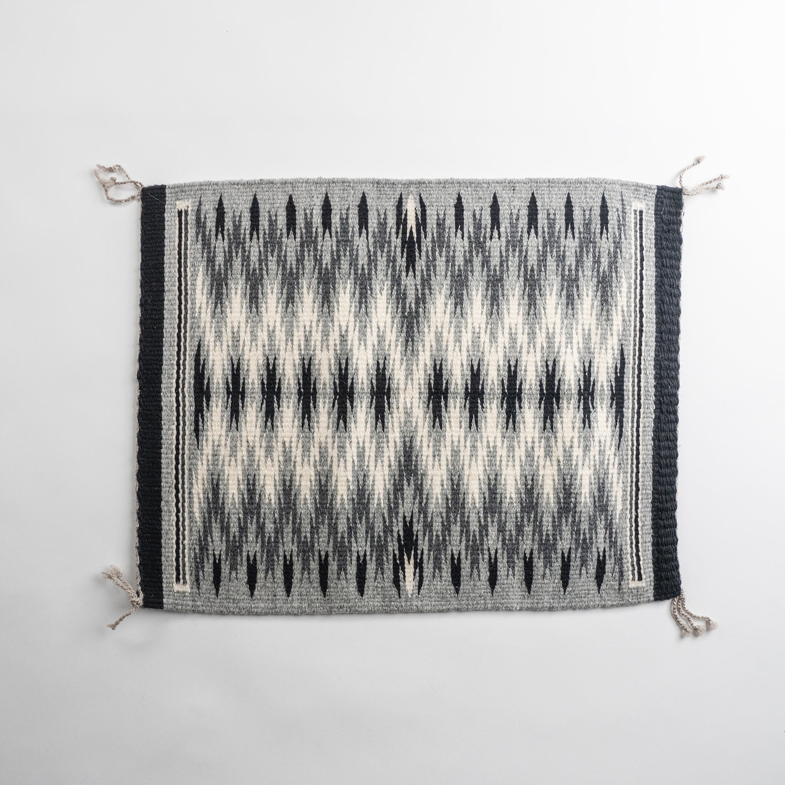 A rectangular woven rug with a geometric Navajo eye dazzler pattern in black, gray, and off-white, featuring black side borders and knotted fringe tassels on the corners.