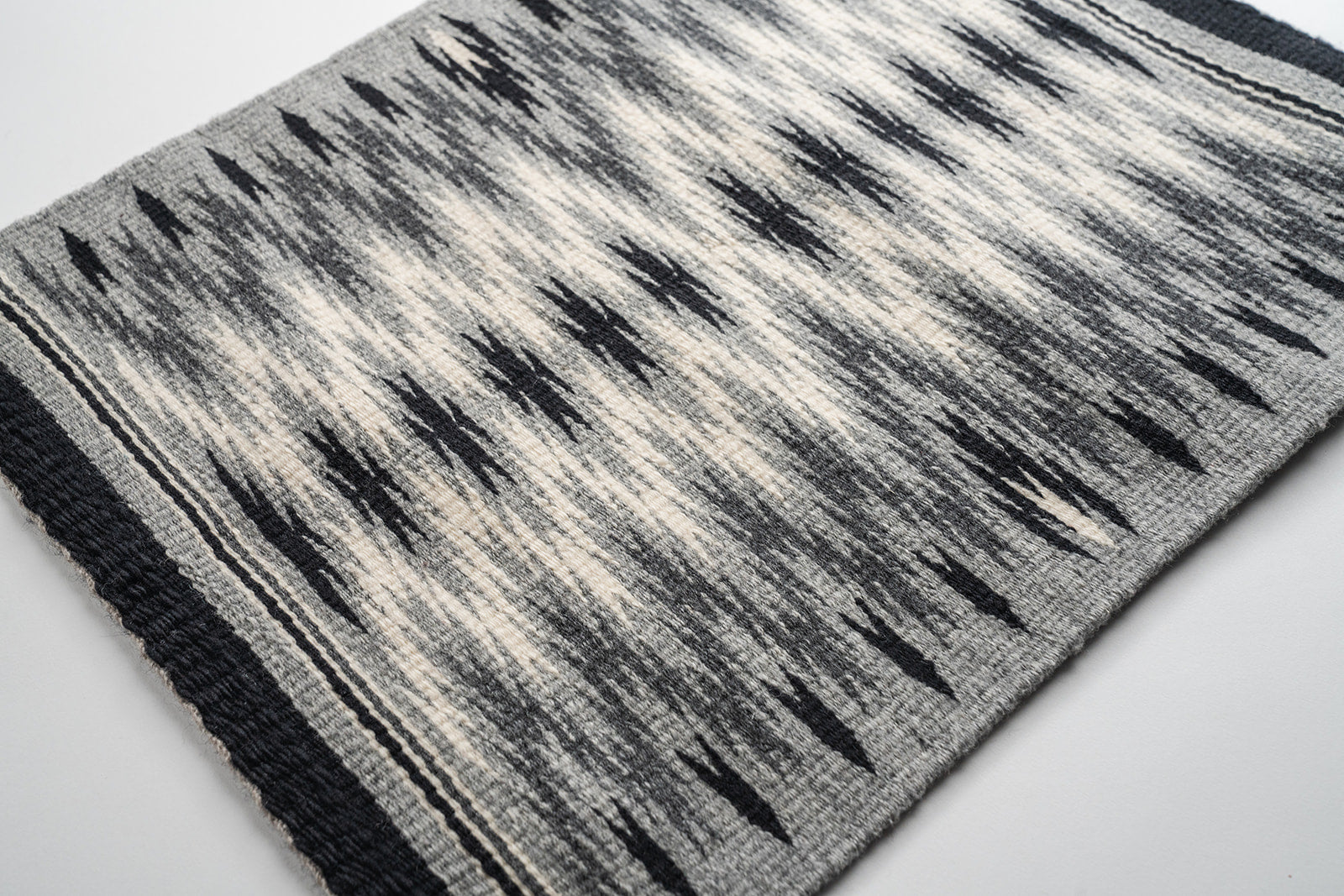 A rectangular woven rug with a geometric Navajo eye dazzler pattern in black, gray, and off-white, featuring black side borders and knotted fringe tassels on the corners.