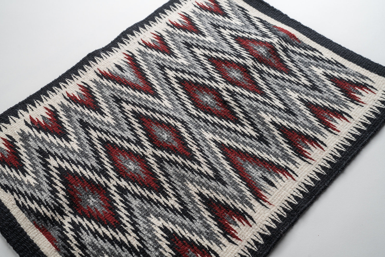 A rectangular woven rug with a Navajo eye dazzler geometric pattern in black, off-white, and dark red, featuring fringed corners.